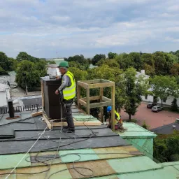 Dwóch dekarzy w kaskach i kamizelkach odblaskowych montuje komin na dachu z blachy miedzianej na tle zabudowań i drzew, widok z góry.