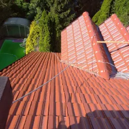 Czerwona dachówka ceramiczna na dachu domu, widok z góry na tle zieleni ogrodowej, z widoczną liną zabezpieczającą i fragmentem rusztowania.