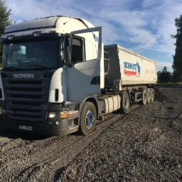 Ciężarówka Scania z otwartymi drzwiami kabiny, zaparkowana na żwirowej drodze, przy częściowo widocznym ogrodzeniu i drzewach w tle.