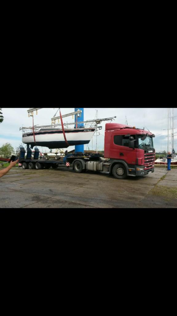 Czerwona ciężarówka Scania przewozi jacht na niskopodwoziowej platformie transportowej, w tle maszty innych jachtów i częściowo widoczna osoba.