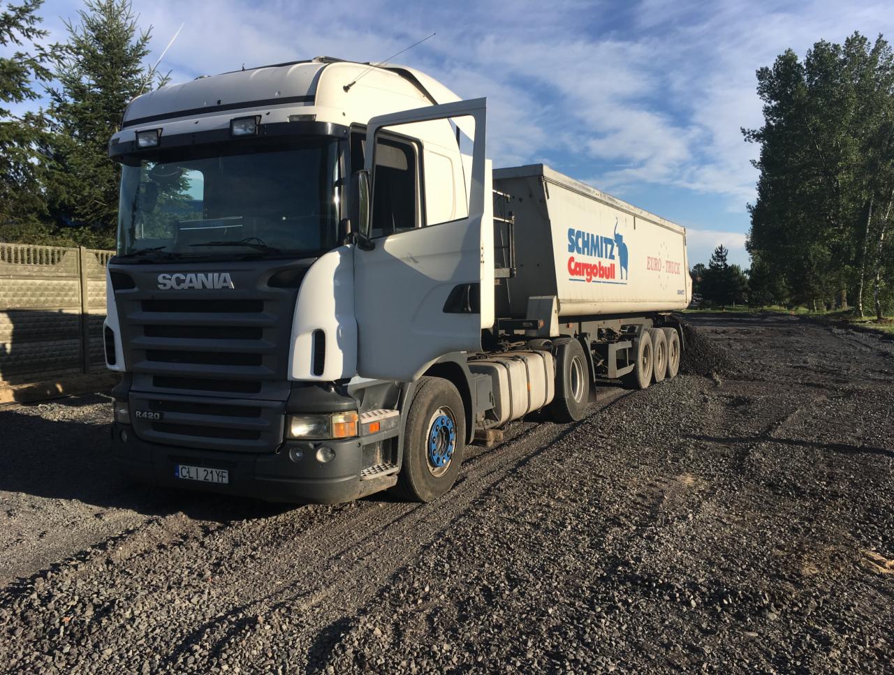 Ciężarówka Scania z otwartymi drzwiami kabiny, zaparkowana na żwirowej drodze, przy częściowo widocznym ogrodzeniu i drzewach w tle.