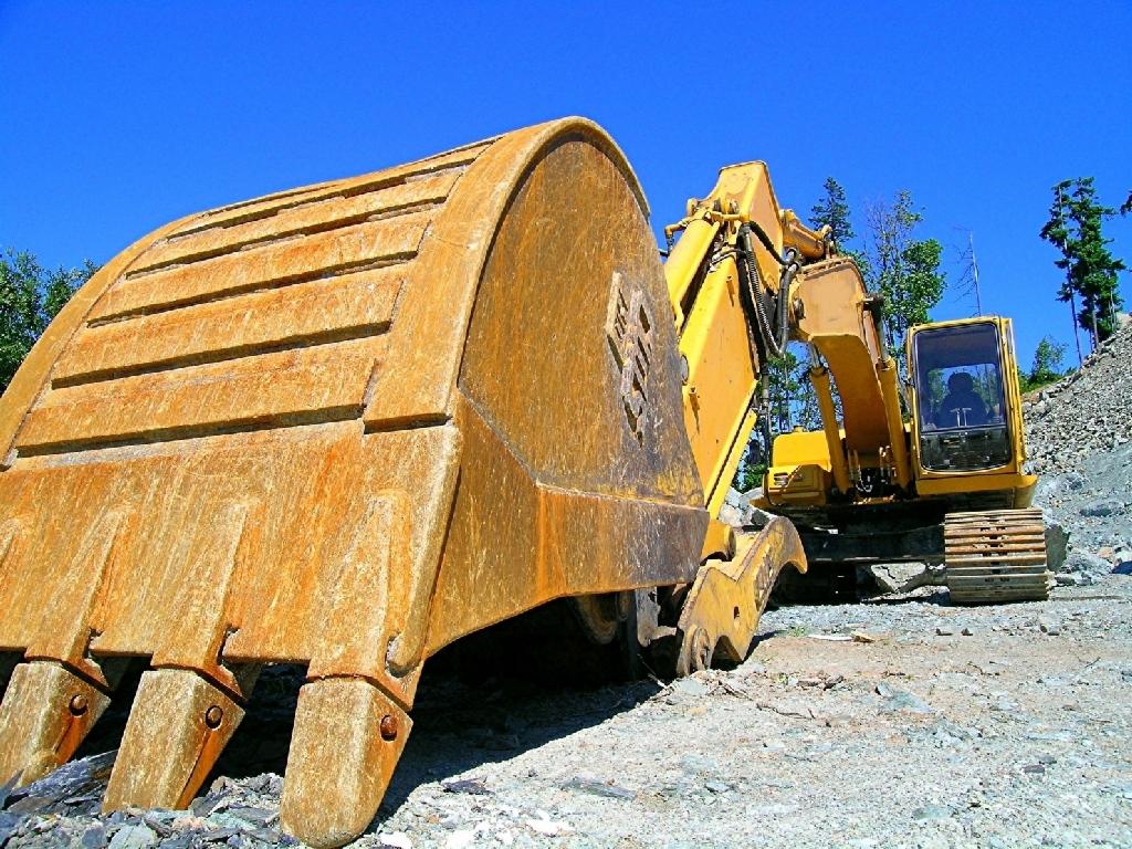 Żółta koparka z dużym, ziemnym łyżką w zbliżeniu na tle błękitnego nieba i skalistego terenu.