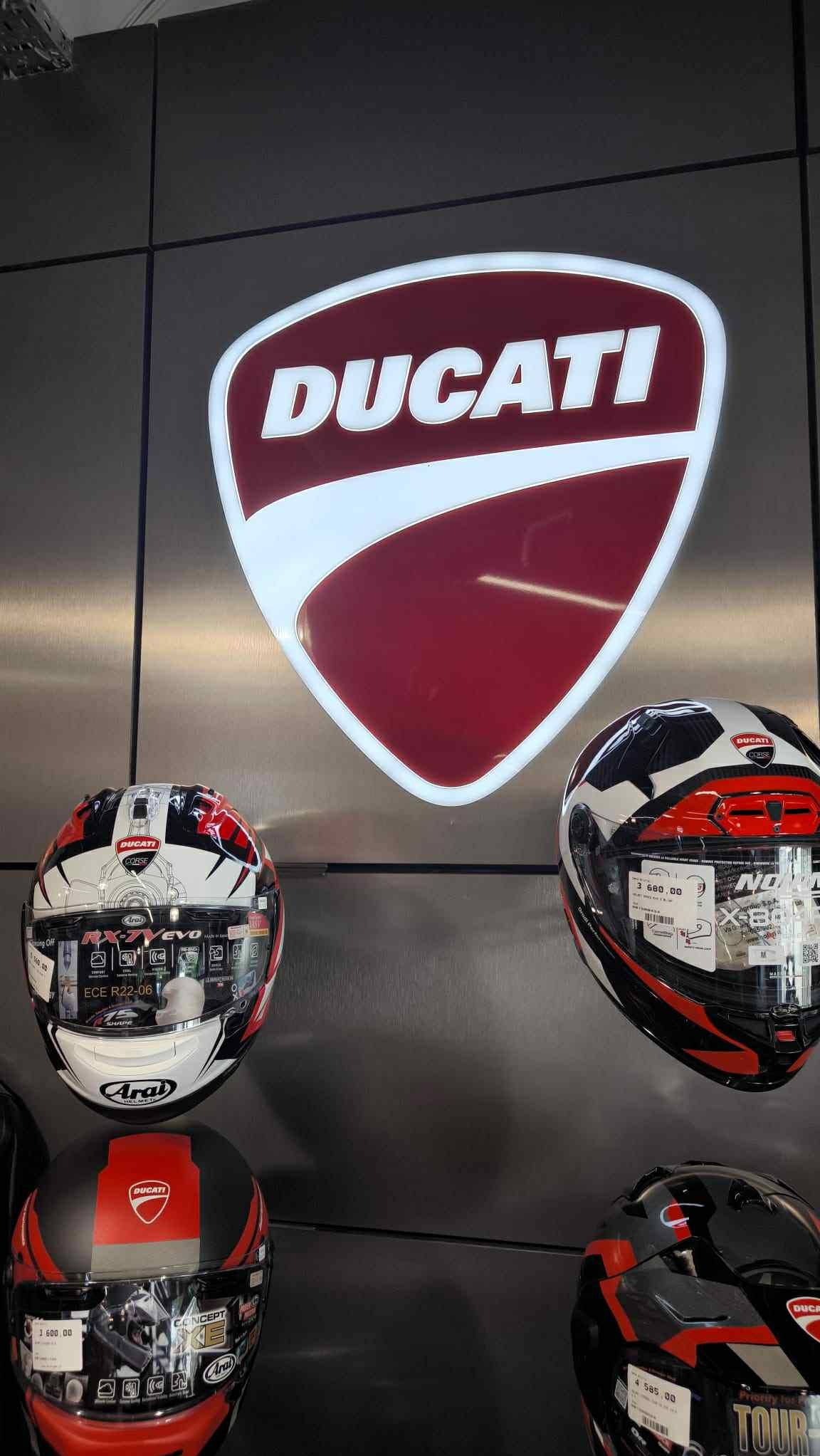 Podświetlany kaseton z logo Ducati na metalowej ścianie, poniżej kaski motocyklowe. Ekspozycja marki w salonie.