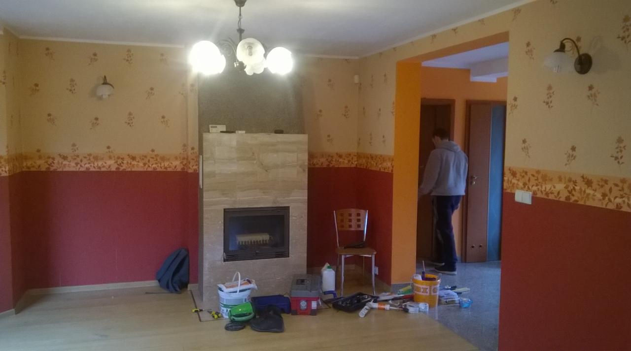 Pokój w trakcie odświeżania z pomalowanymi ścianami na bordowo i beżowo z kwiatowym motywem, widocznym kominkiem obłożonym płytkami imitującymi kamień, narzędziami malarskimi i osobą w tle.
