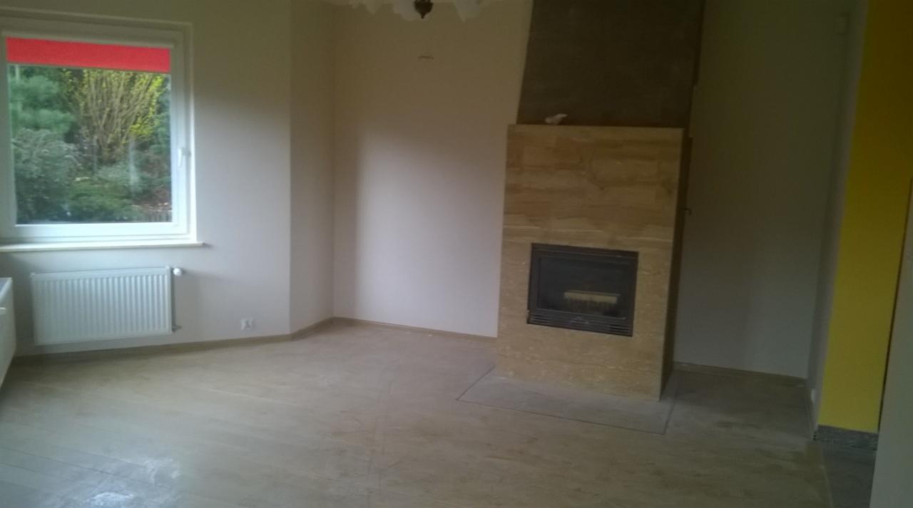 Wnętrze pokoju z kominkiem obłożonym trawertynem, podłoga wyłożona panelami, widoczne ślady po szpachlowaniu na ścianach i suficie, okno z czerwonym roletą.