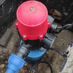 Stacje uzdatniania wody - Zbliżenie na instalację w czarnej skrzynce: czerwony zawór z czterema śrubami, niebieska rura oraz czarne przewody, otoczone żwirem.