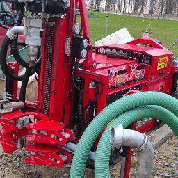 Stacje uzdatniania wody - Czerwona wiertnica do studni głębinowych z zielonym wężem ssawnym na placu budowy, w tle quad i czerwony bus.