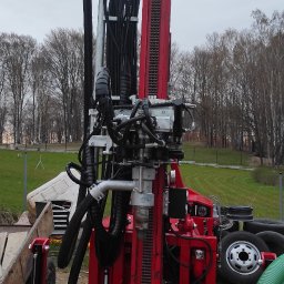 Stacje uzdatniania wody - Czerwona wiertnica do studni głębinowych na placu budowy, widoczne elementy hydrauliczne, przewody, i teren z drzewami w tle.