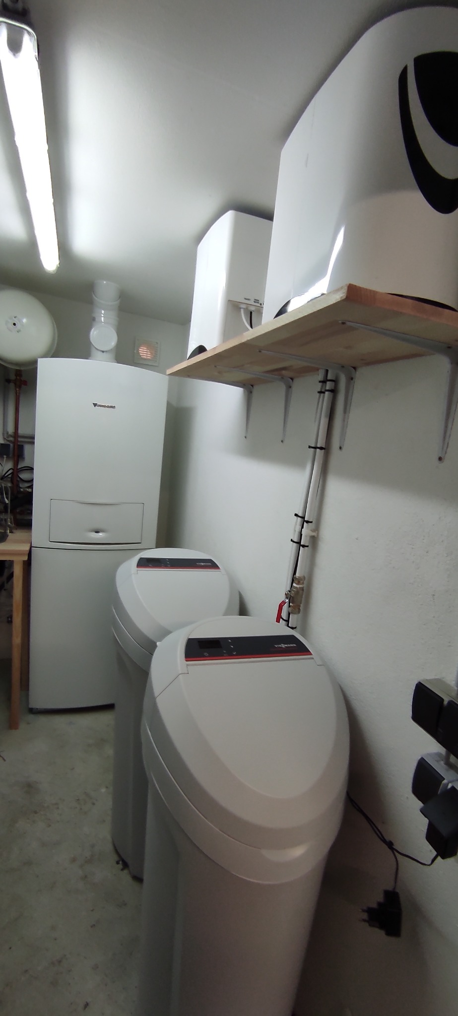 Dwa białe filtry do wody Viessmann stoją obok pieca grzewczego w pomieszczeniu technicznym z betonową podłogą, oświetlonym jarzeniówką, z widoczną półką na ścianie.