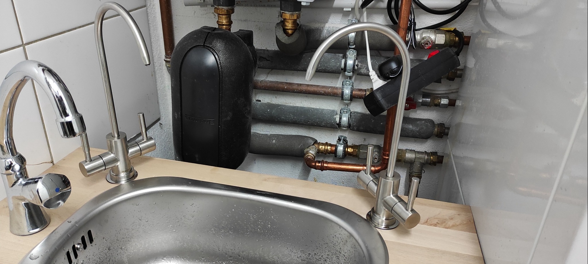 Montaż armatury kuchennej: trzy baterie, w tym jedna z filtrem wody, zainstalowane przy zlewie ze stali nierdzewnej. W tle widoczne rury instalacyjne i czarny zbiornik. Ujęcie z bliska, ukazujące...