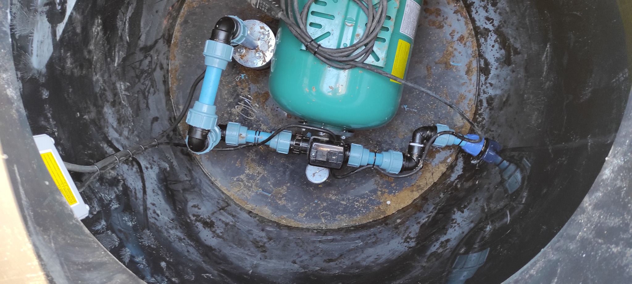 Wnętrze czarnej studni z zamontowaną zieloną pompą hydroforową, niebieskimi rurami PCV i manometrem, widoczne z góry.