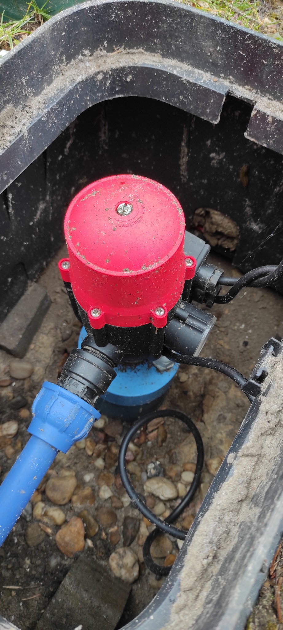 Zbliżenie na instalację w czarnej skrzynce: czerwony zawór z czterema śrubami, niebieska rura oraz czarne przewody, otoczone żwirem.