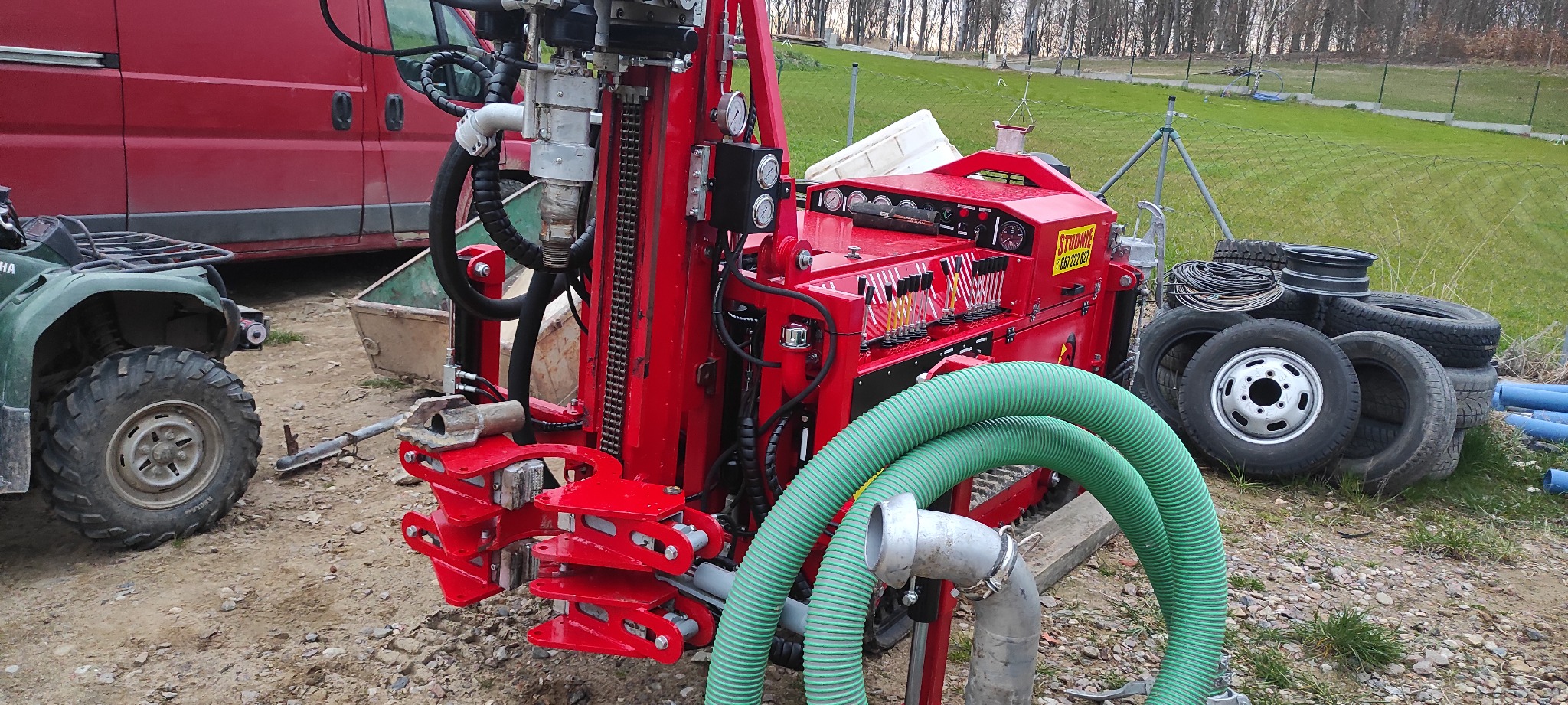 Czerwona wiertnica do studni głębinowych z zielonym wężem ssawnym na placu budowy, w tle quad i czerwony bus.