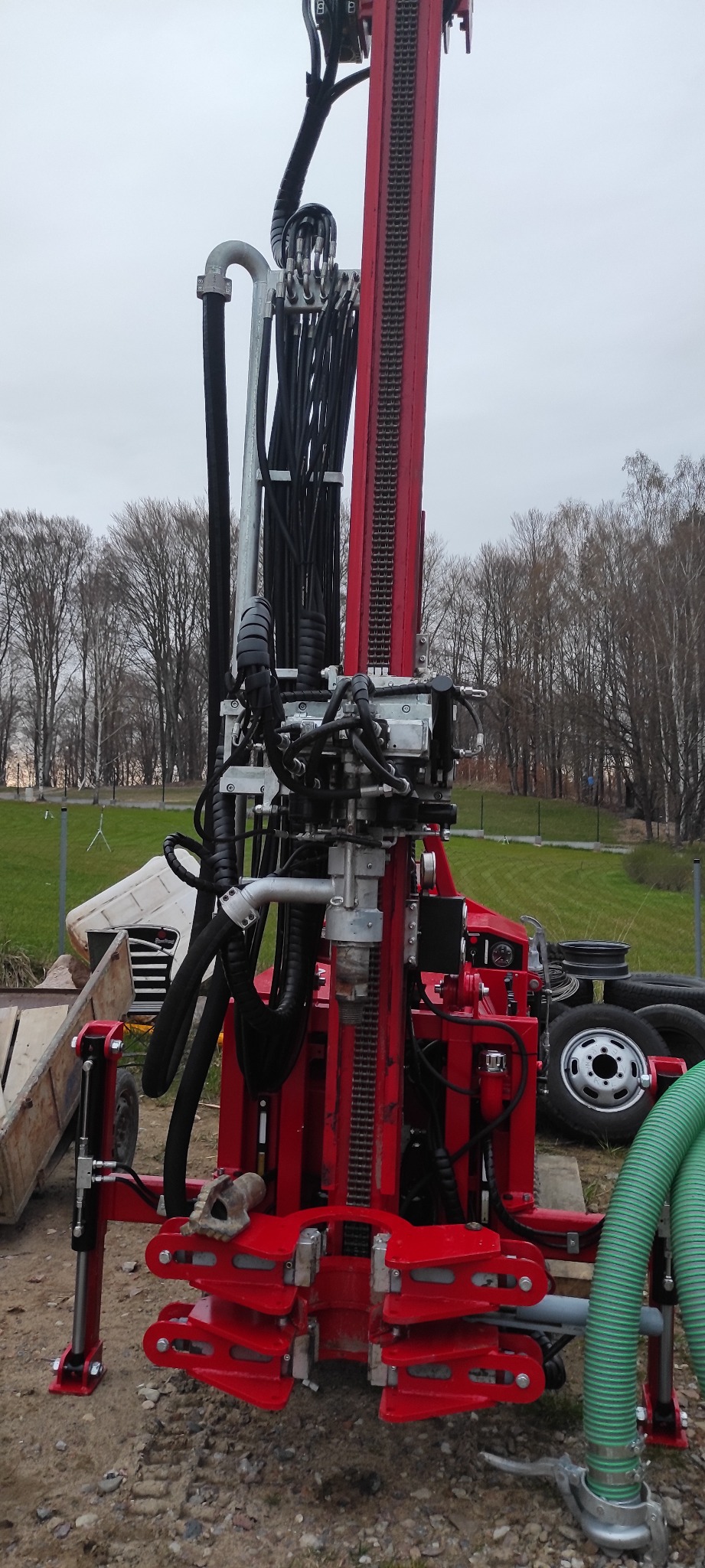 Czerwona wiertnica do studni głębinowych na placu budowy, widoczne elementy hydrauliczne, przewody, i teren z drzewami w tle.