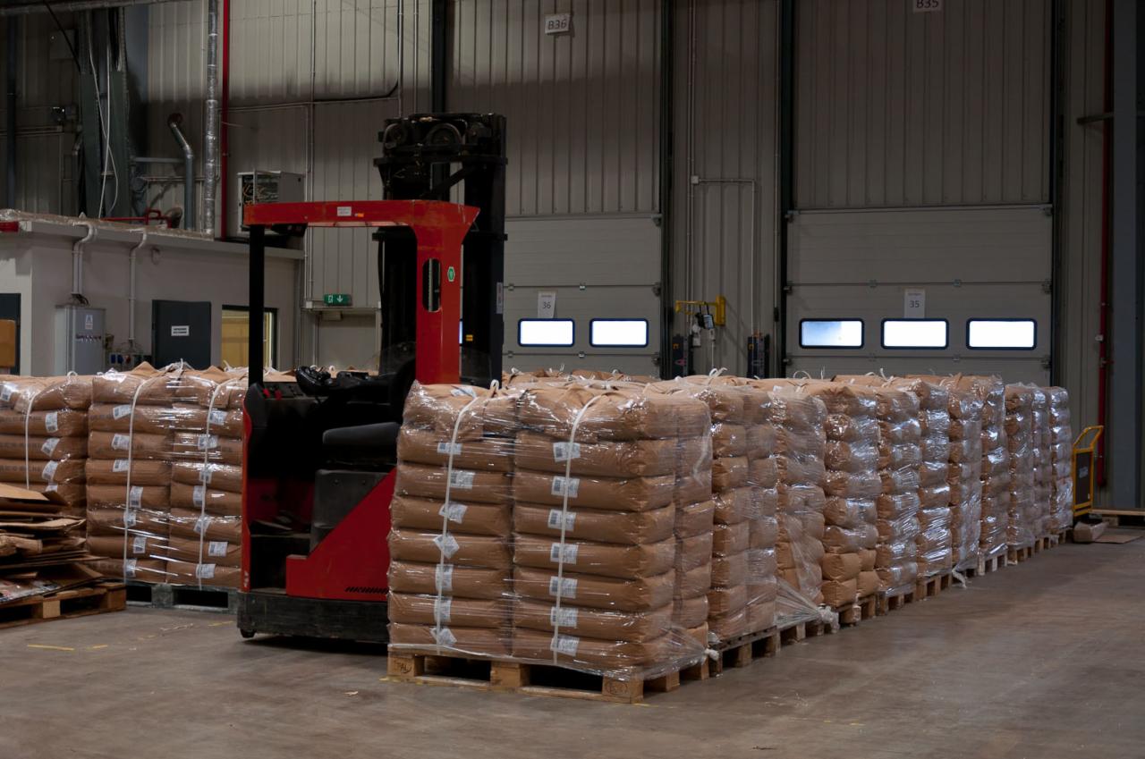 Czerwony wózek widłowy między paletami z workami w magazynie, owiniętymi folią stretch, widoczne bramy magazynowe z oknami.