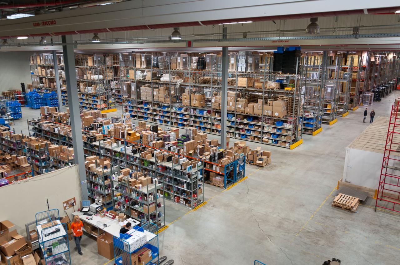 Szeroki kadr z lotu ptaka na duży magazyn z wysokimi regałami wypełnionymi kartonami i różnymi produktami, z pracownikami przy stanowisku pakowania po lewej stronie i dwoma osobami spacerującymi...