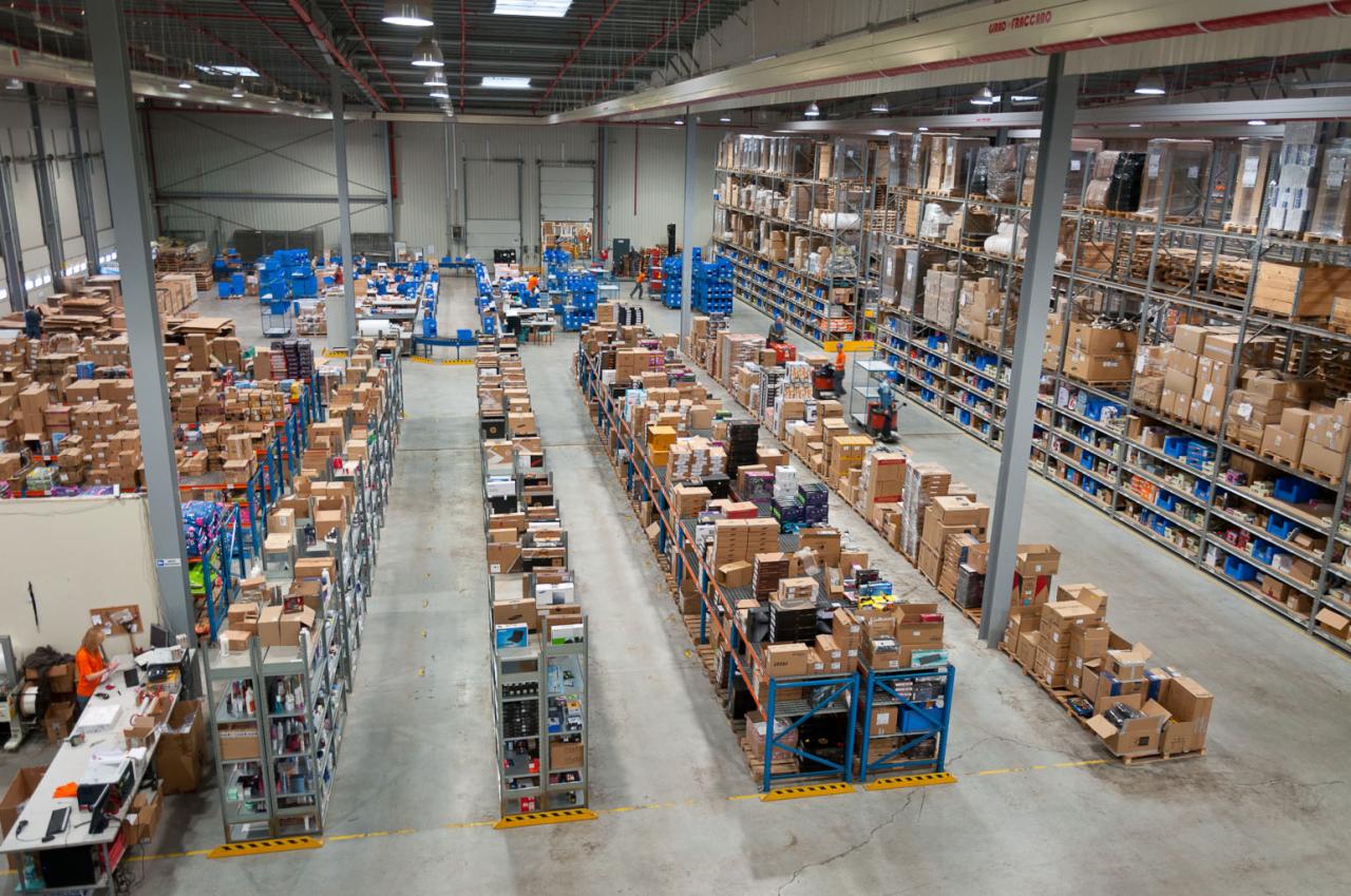 Przestronny magazyn wysokiego składowania z regałami wypełnionymi kartonami i różnorodnymi towarami, widok z góry, pracownicy i wózki widłowe w oddali.