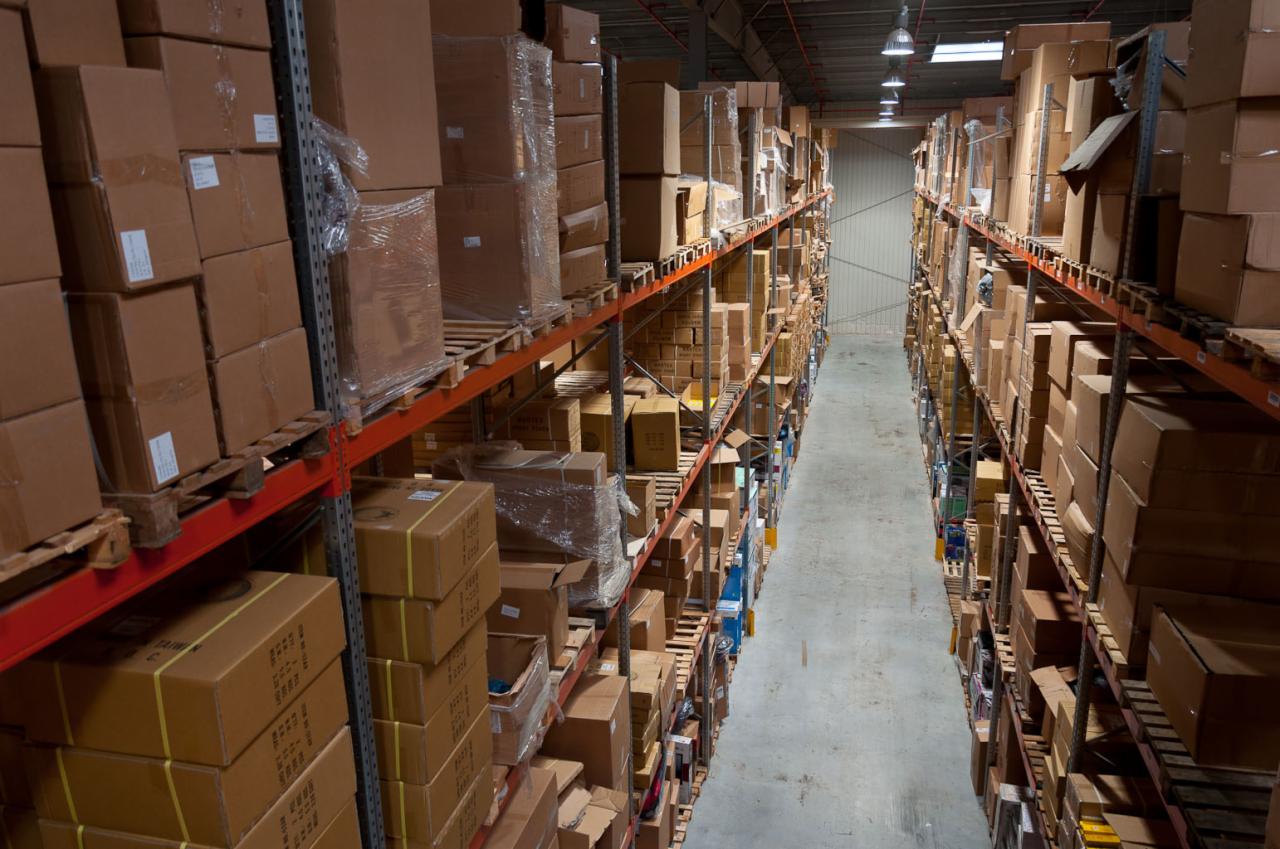 Długi korytarz magazynowy z wysokimi regałami wypełnionymi kartonami, niektóre owinięte folią, oświetlony przez lampy sufitowe.