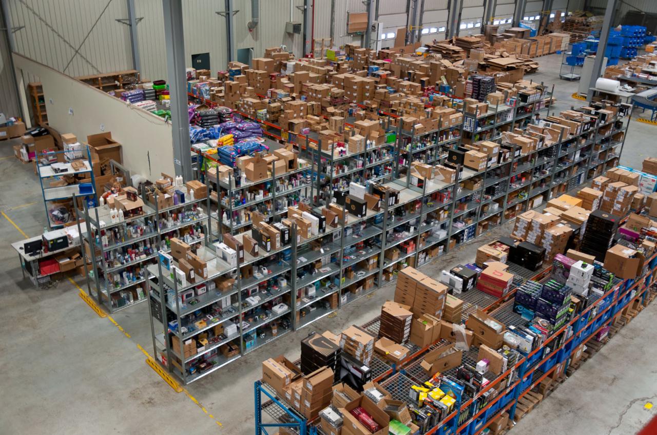 Widok z góry na magazyn z metalowymi regałami wypełnionymi kartonami, pudełkami i towarem różnego rodzaju, w tle widoczne palety i kartony składowane luzem.
