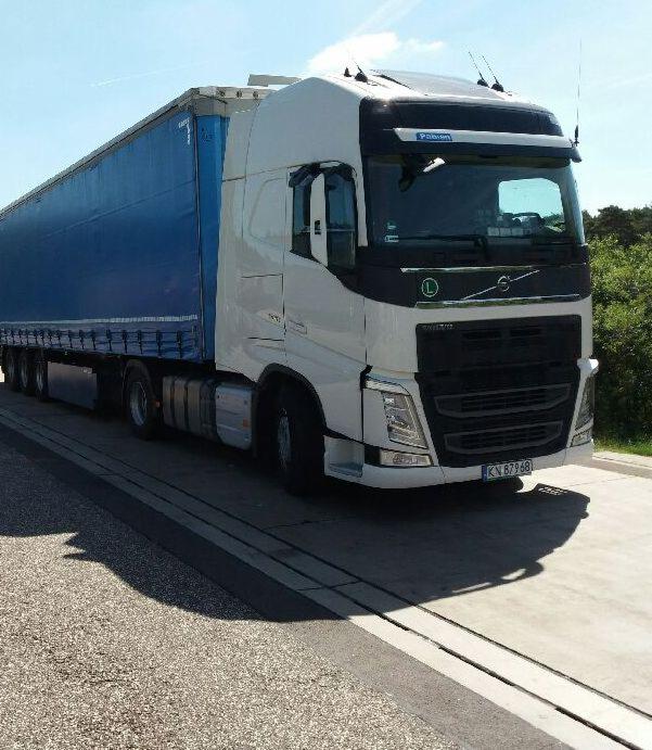 Biała ciężarówka Volvo z niebieską naczepą na betonowej platformie w słoneczny dzień, widoczne tablice rejestracyjne.