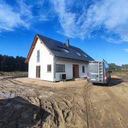 Ireneusz Kremin - Biały dom z ciemną dachówką w trakcie budowy, z widocznym samochodem dostawczym na błotnistym terenie. Błękitne niebo z chmurami w tle. Widoczne okna i drzwi z drewnianymi ramami.