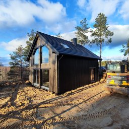 Ireneusz Kremin - Nowoczesny, ciemny dom szkieletowy z dużymi oknami, na piaszczystym terenie z widoczną koparką. Błękitne niebo z chmurami w tle. Charakterystyczny kształt okna.