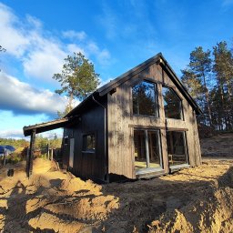 Ireneusz Kremin - Nowoczesny dom z ciemnego drewna z dużymi oknami, w otoczeniu piaszczystego terenu i drzew. Widok z dołu podkreśla bryłę budynku i błękitne niebo.