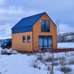 Ireneusz Kremin - Nowoczesny, drewniany dom z niebieskim dachem w zimowej scenerii. Widoczny częściowo taras i samochód dostawczy. Krajobraz za domem pokryty śniegiem.