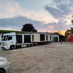 Ireneusz Kremin - Transport domu modułowego na lawecie ciężarówki na tle budynku z zielonymi pnączami i zachodzącego słońca. Widoczny fragment samochodu osobowego.