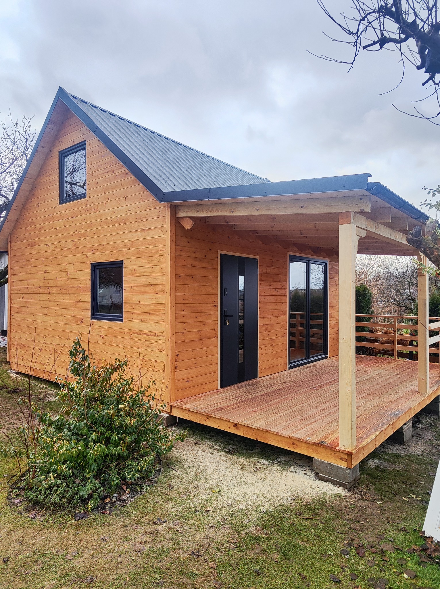 Nowoczesny dom szkieletowy z drewnianą elewacją i tarasem, czarne okna i drzwi, metalowy dach, widok z zewnątrz w pochmurny dzień. Elementy wykończeniowe.