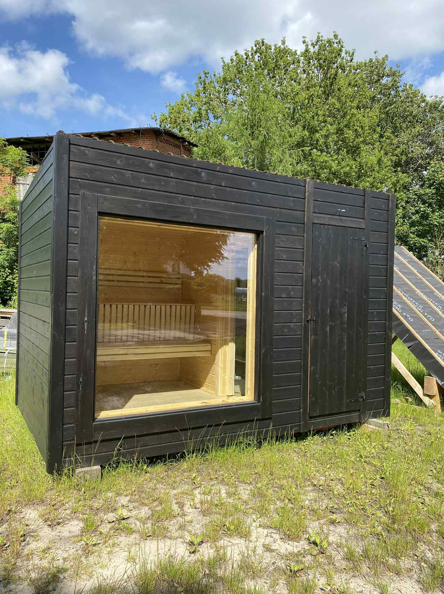Czarna, drewniana sauna ogrodowa z dużym oknem, przez które widać drewniane ławki wewnątrz. Obok widoczna mała, wąska furtka. Całość na trawiastym podłożu.