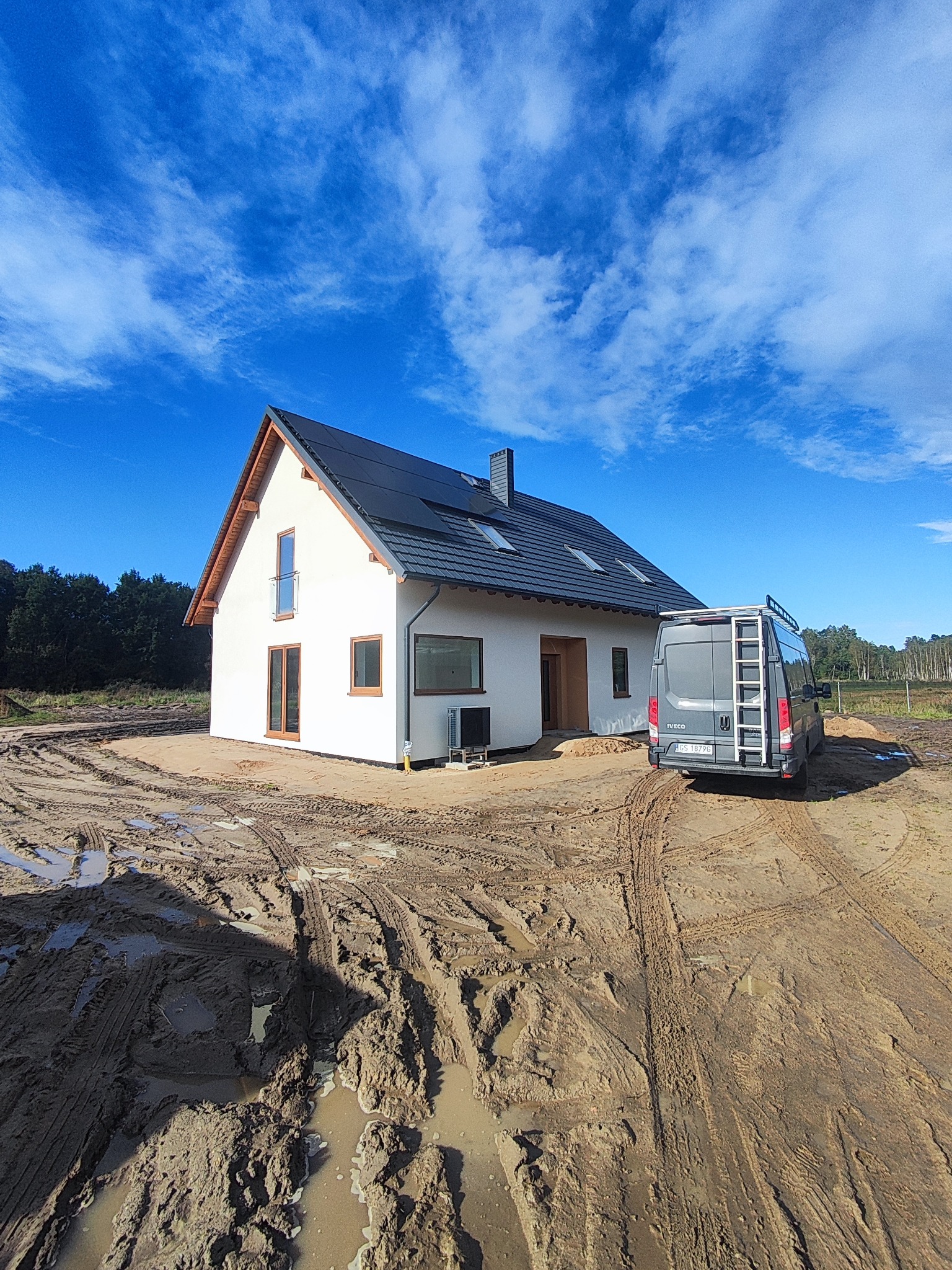 Biały dom z ciemną dachówką w trakcie budowy, z widocznym samochodem dostawczym na błotnistym terenie. Błękitne niebo z chmurami w tle. Widoczne okna i drzwi z drewnianymi ramami.