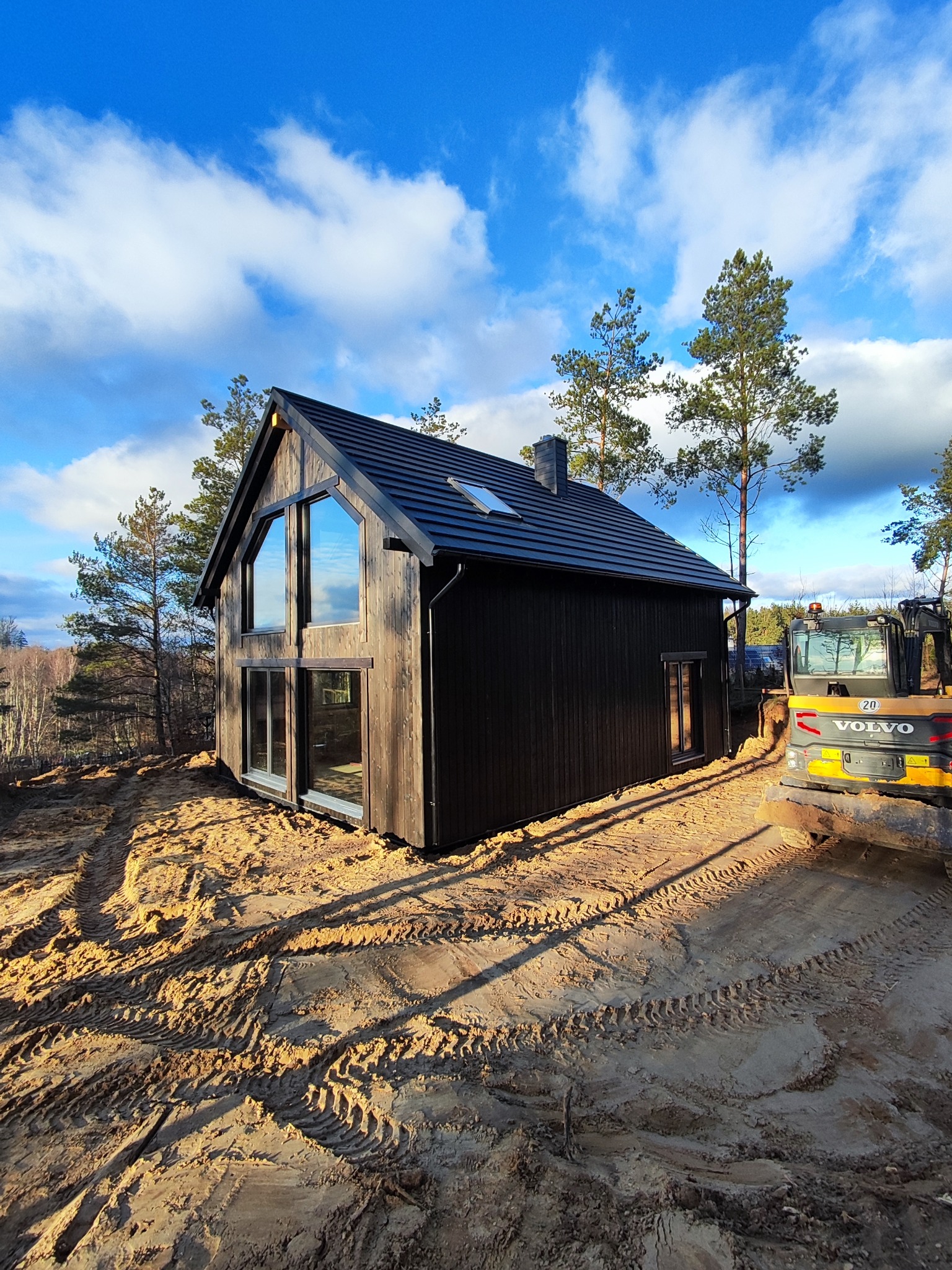 Nowoczesny, ciemny dom szkieletowy z dużymi oknami, na piaszczystym terenie z widoczną koparką. Błękitne niebo z chmurami w tle. Charakterystyczny kształt okna.