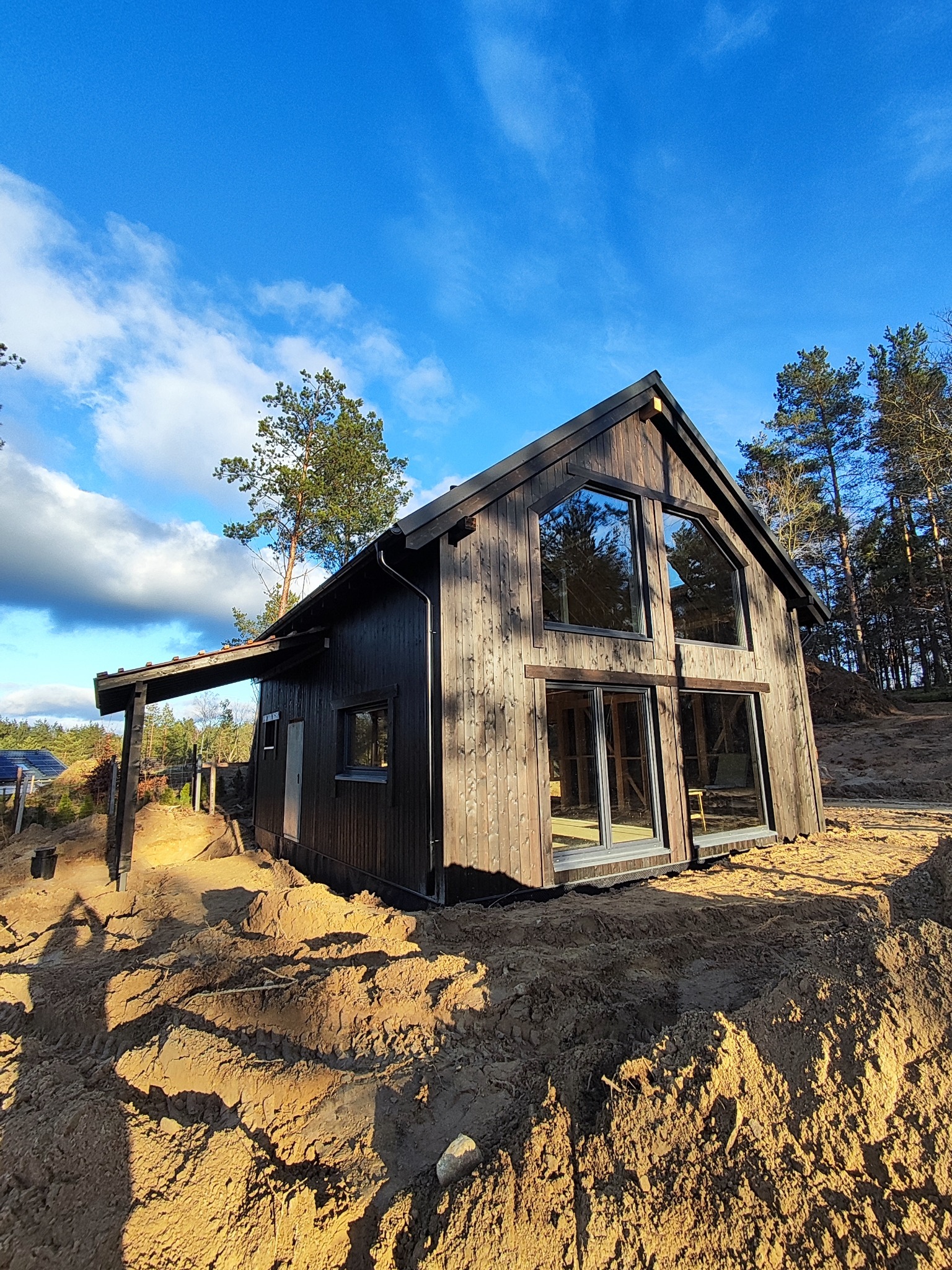 Nowoczesny dom z ciemnego drewna z dużymi oknami, w otoczeniu piaszczystego terenu i drzew. Widok z dołu podkreśla bryłę budynku i błękitne niebo.
