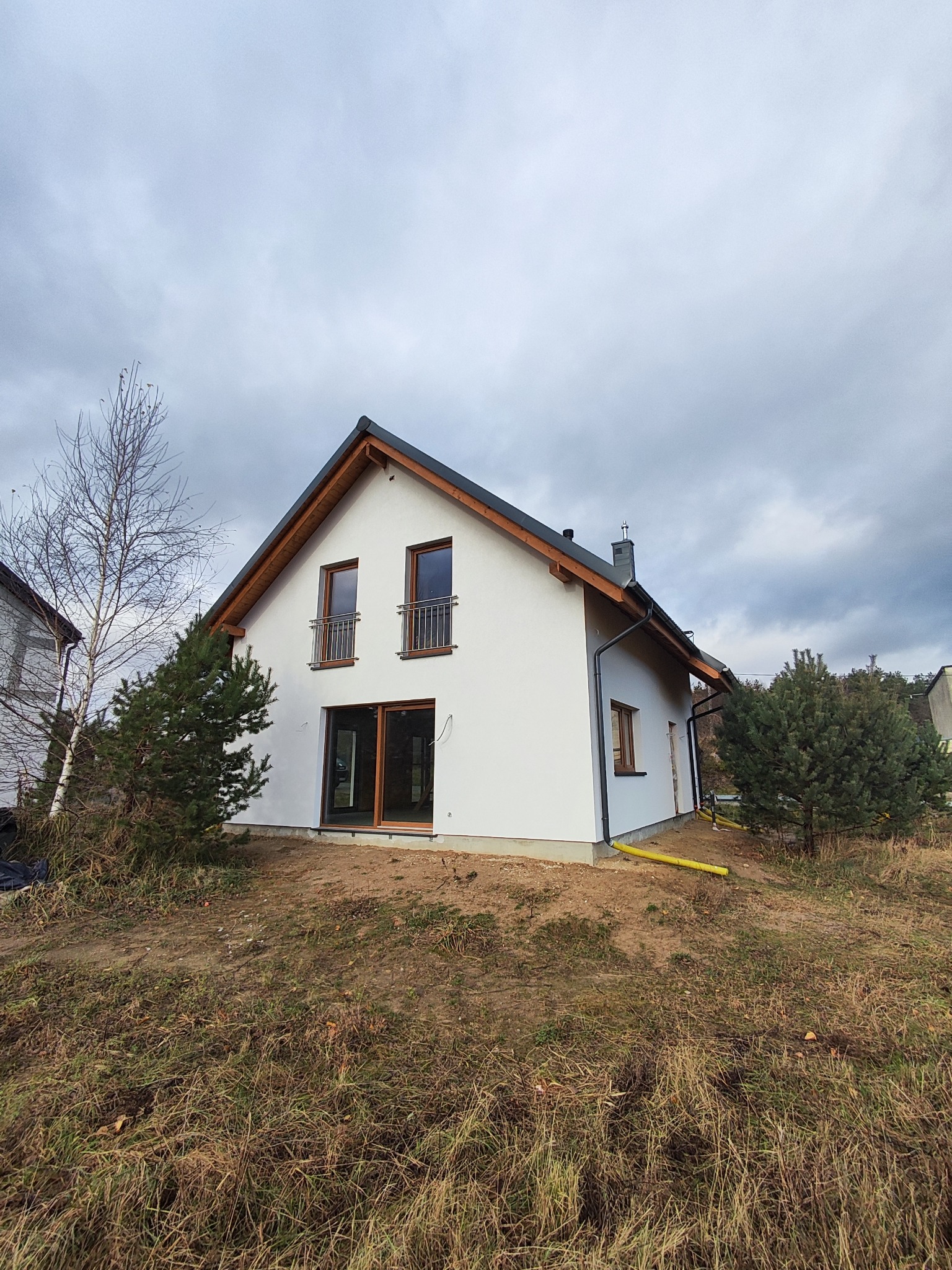 Biały dom jednorodzinny z ciemną dachówką i drewnianymi elementami wykończeniowymi, widoczny z perspektywy ogrodu w pochmurny dzień. Dom na wzniesieniu.