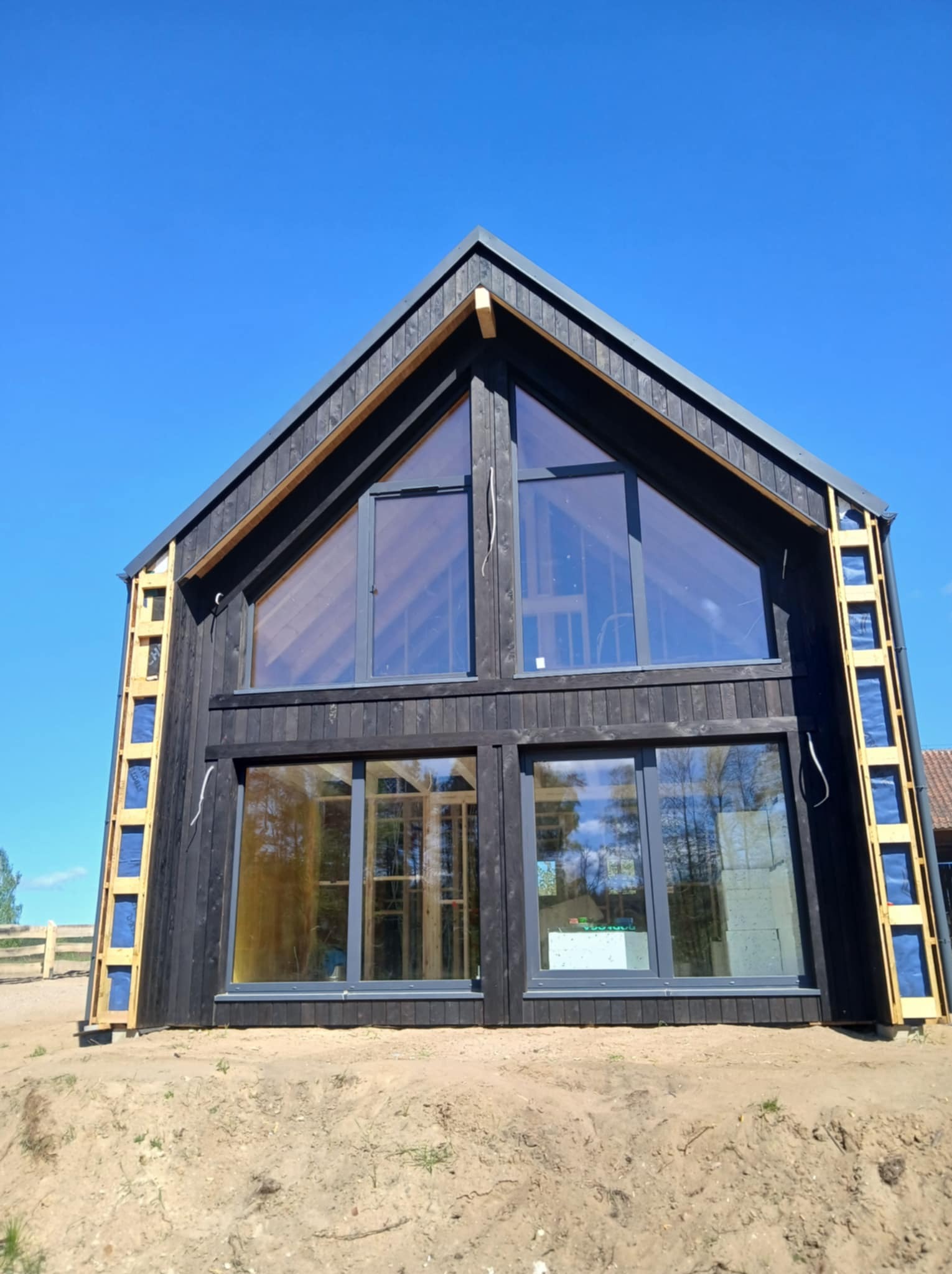 Ciemny, nowoczesny dom z dużymi oknami w trakcie budowy, widoczne elementy konstrukcyjne i ocieplenie. Błękitne niebo w tle.