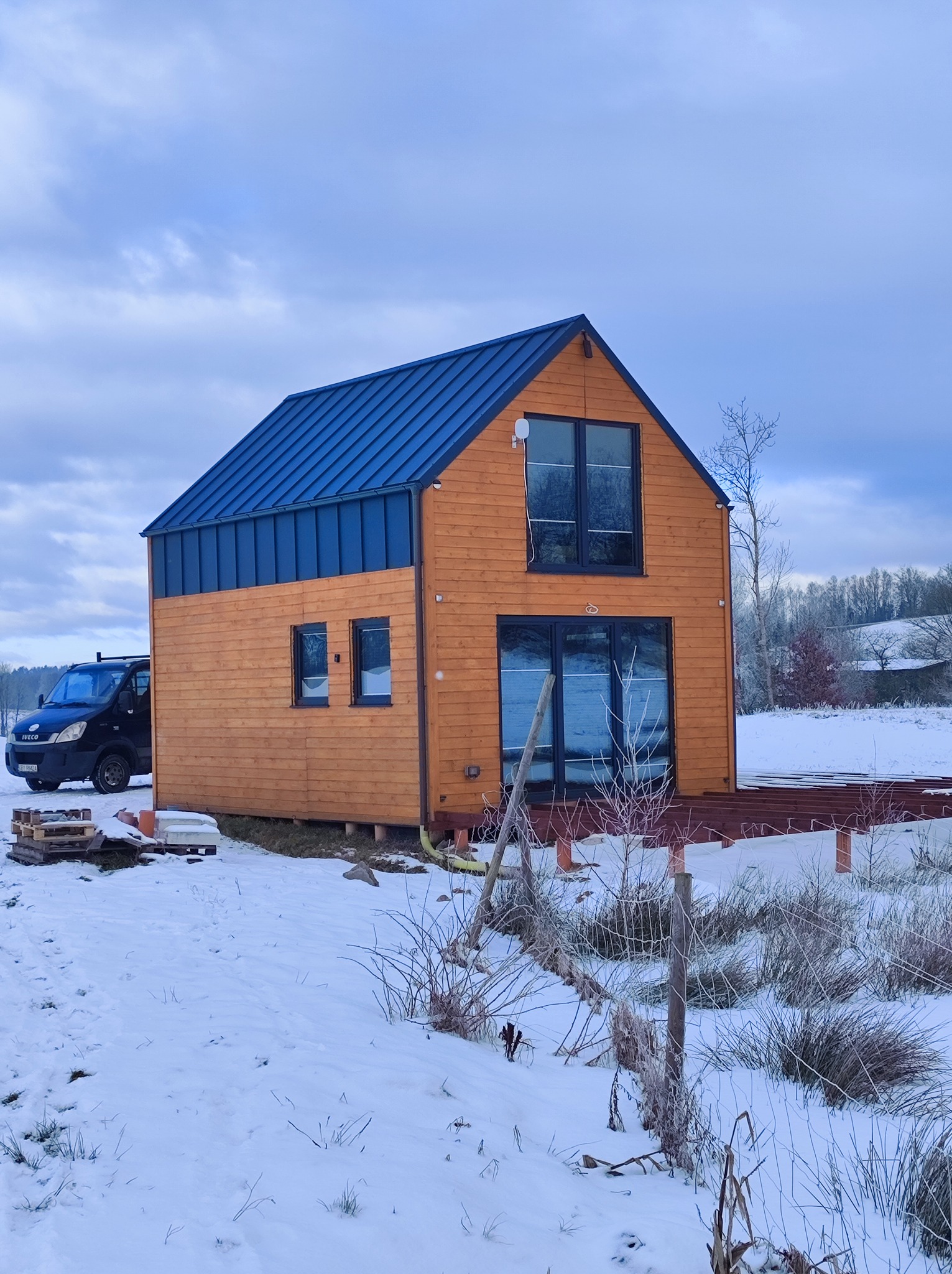 Nowoczesny, drewniany dom z niebieskim dachem w zimowej scenerii. Widoczny częściowo taras i samochód dostawczy. Krajobraz za domem pokryty śniegiem.