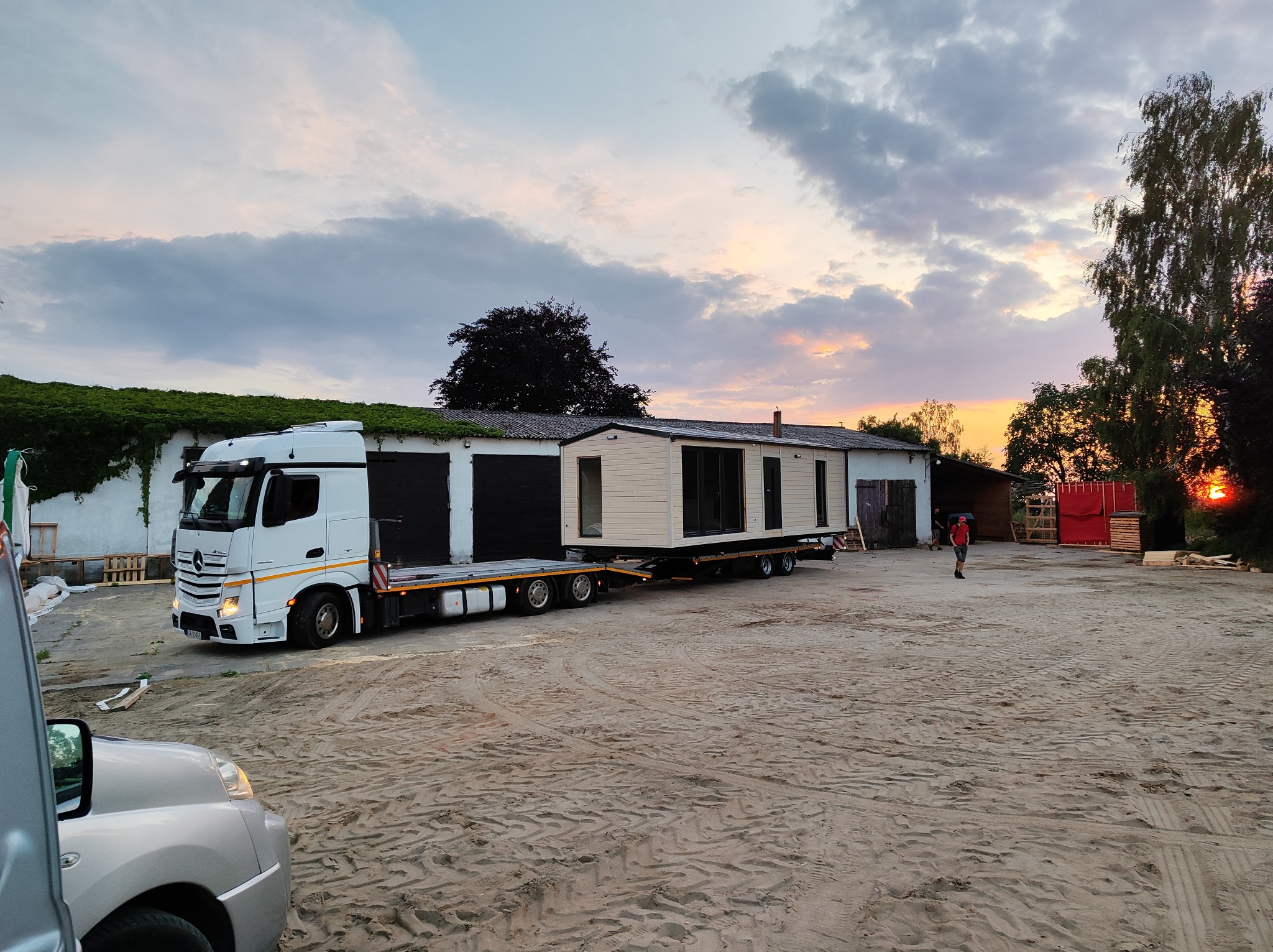Transport domu modułowego na lawecie ciężarówki na tle budynku z zielonymi pnączami i zachodzącego słońca. Widoczny fragment samochodu osobowego.