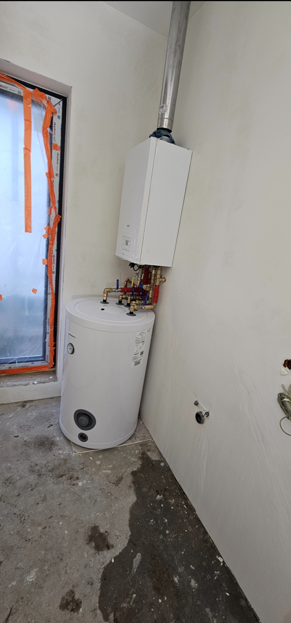 Biały, wiszący piec gazowy z podłączonym zasobnikiem wody, widoczne rury miedziane z izolacją, zamontowane w surowym wnętrzu, okno zabezpieczone folią.