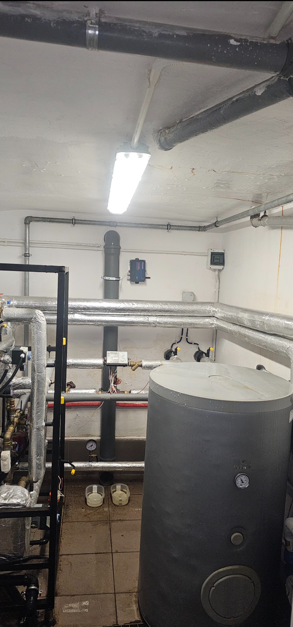 Kotłownia z szarym zbiornikiem na wodę, rurami w srebrnej izolacji, czarną konstrukcją wsporczą i dwoma plastikowymi pojemnikami na podłodze.