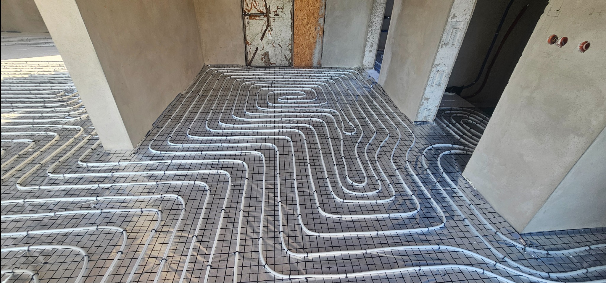 Rozłożone rury ogrzewania podłogowego na siatce w surowym wnętrzu, widoczne ściany w stanie deweloperskim i otwory na instalacje elektryczne.