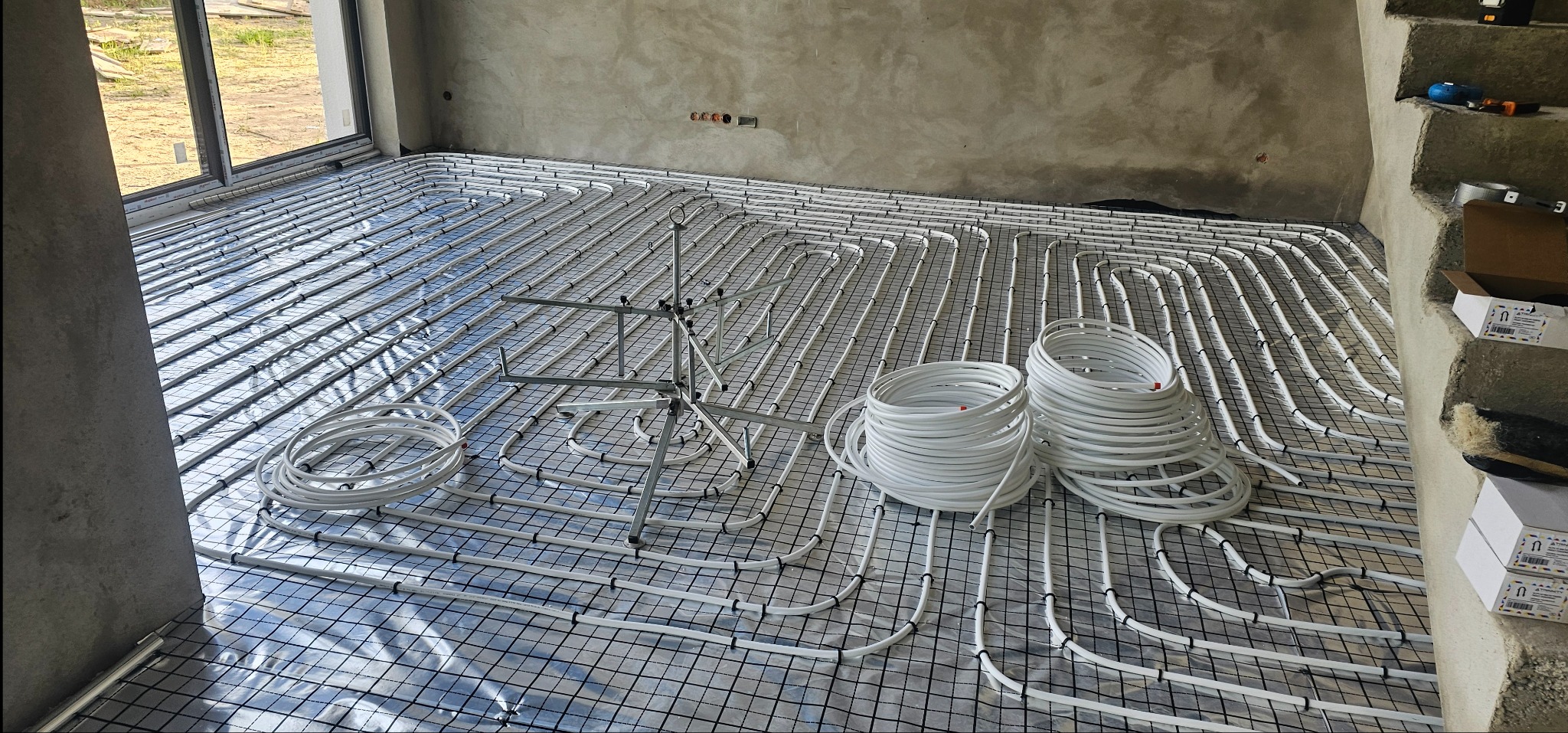 Rozłożona instalacja ogrzewania podłogowego z rurami na folii z siatką, widoczne zwoje rur i statyw montażowy w surowym wnętrzu.