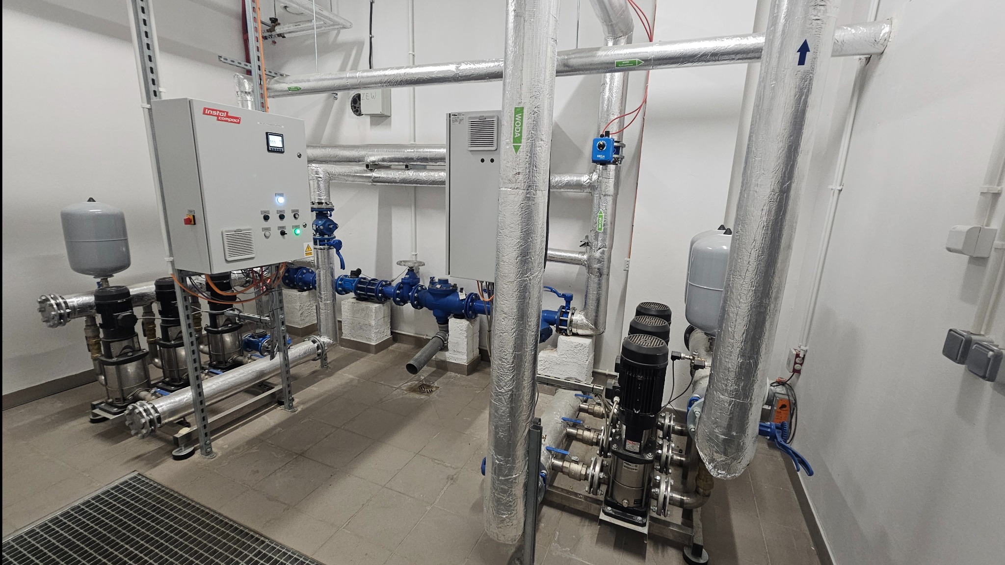Węzeł hydroforowy z dwoma zestawami pompowymi, zbiornikami przeponowymi i szafami sterowniczymi, rury w izolacji aluminiowej z oznaczeniami kierunku przepływu, kratka ściekowa w tle.