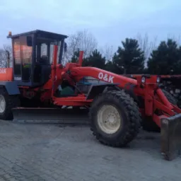 Czerwony równiarka O&K F156A zaparkowany na betonowej nawierzchni, widoczne drzewa w tle.