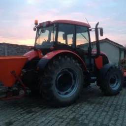Czerwony traktor z pomarańczowym rozsiewaczem na kostce brukowej, w tle betonowe ogrodzenie i budynek gospodarczy, niebo o zmierzchu.