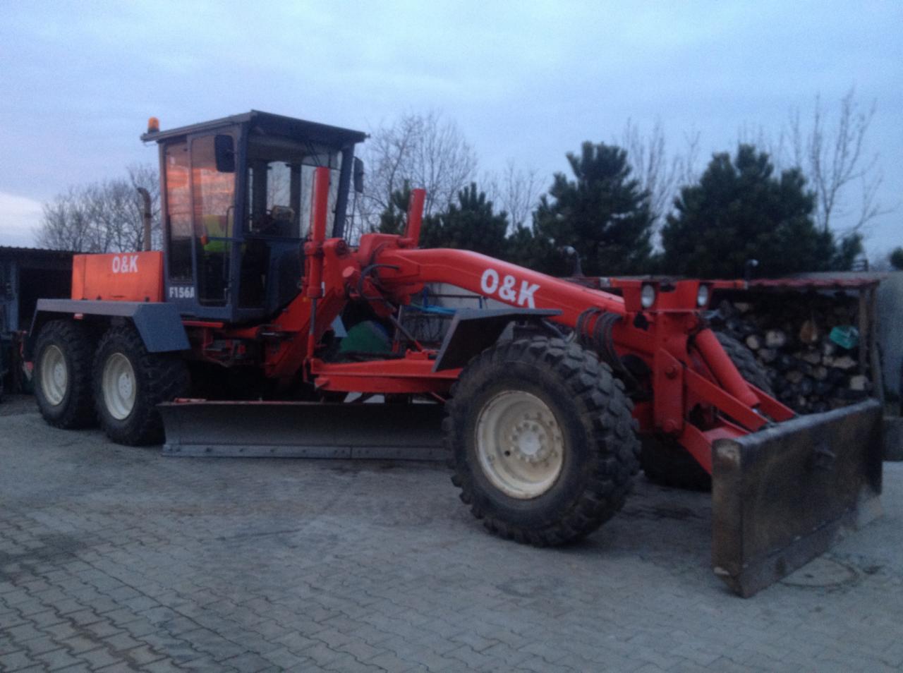 Czerwony równiarka O&K F156A zaparkowany na betonowej nawierzchni, widoczne drzewa w tle.