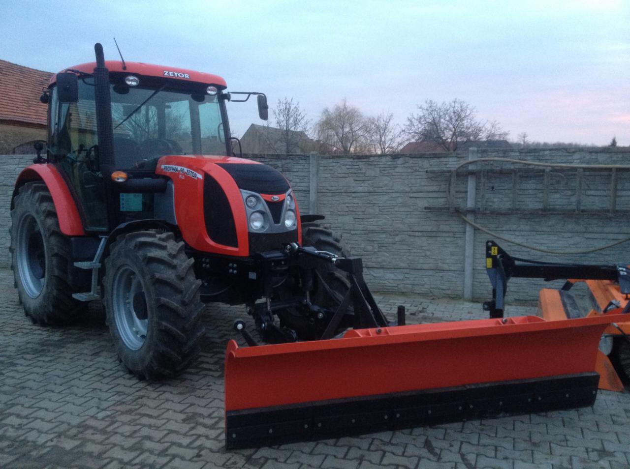 Czerwony traktor Zetor z zamontowanym pługiem do śniegu, stojący na kostce brukowej przed betonowym ogrodzeniem.