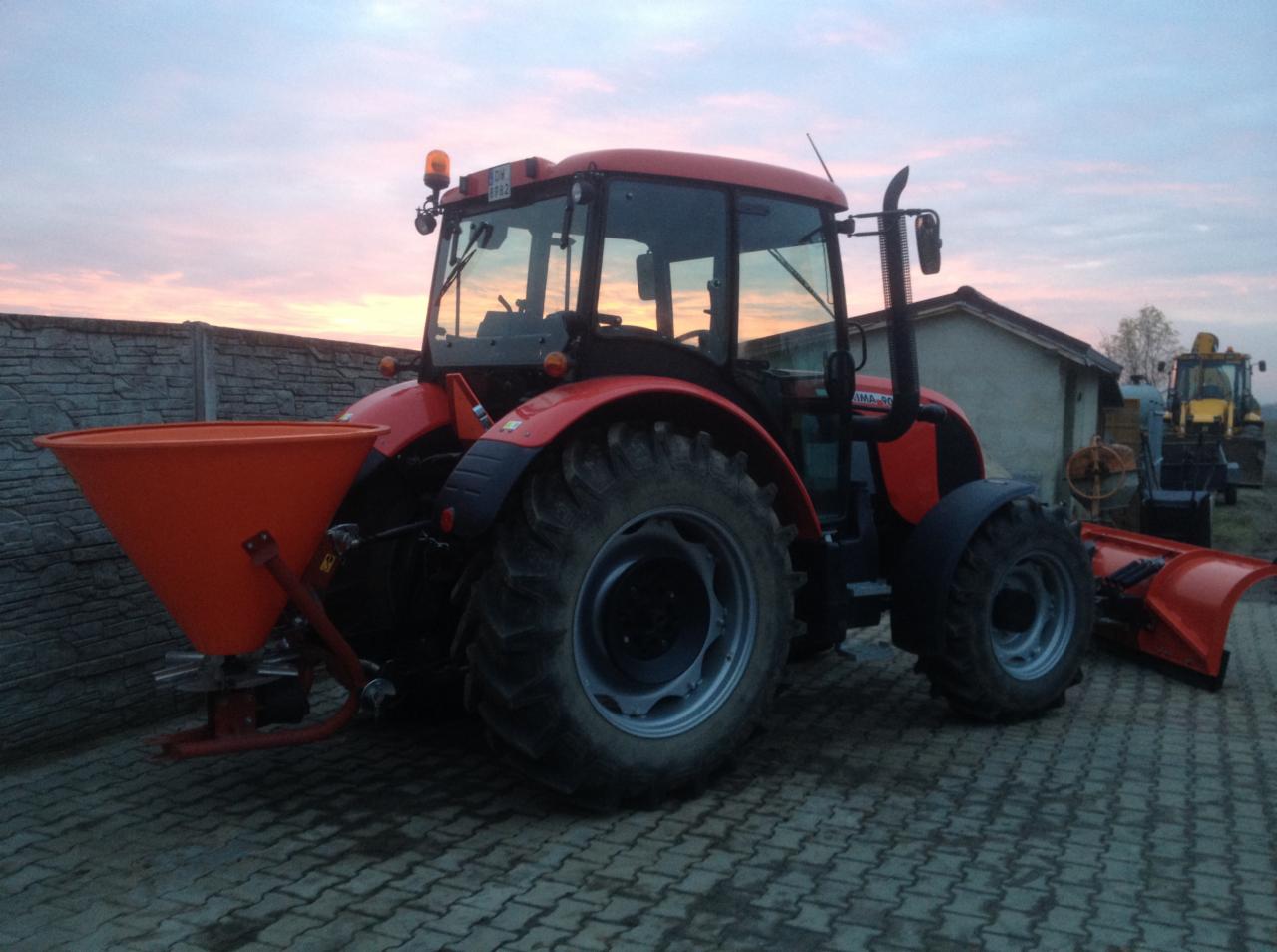 Czerwony traktor z pomarańczowym rozsiewaczem na kostce brukowej, w tle betonowe ogrodzenie i budynek gospodarczy, niebo o zmierzchu.