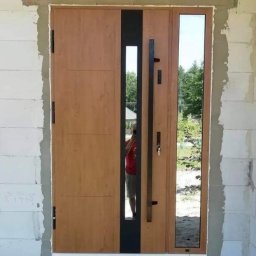 FHU TOMEX Tomasz Słoniewski - Nowoczesne drzwi zewnętrzne z drewnianym wykończeniem, czarnym akcentem i długim uchwytem, zamontowane w surowym otoczeniu budowlanym w Makowie Mazowieckim, z odbiciem fotografa w szybie.