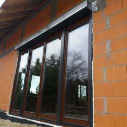FHU TOMEX Tomasz Słoniewski - Czterodzielne okno z brązową ramą i szarą roletą zewnętrzną zamontowane w surowej, ceglanej ścianie budynku w trakcie budowy.